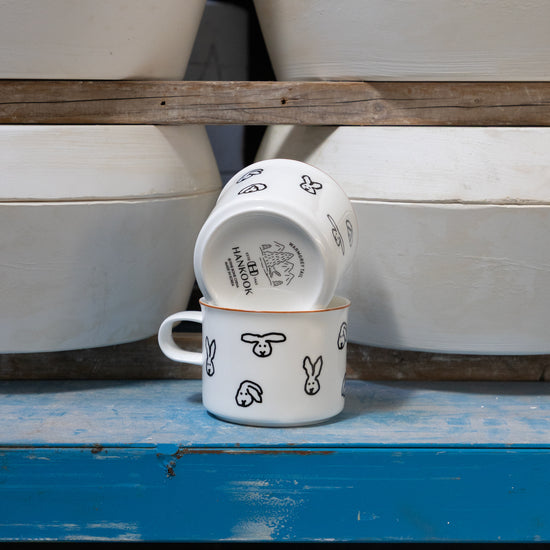 Two white ceramic cups with black designs on a blue surface, with stacked white ceramic pots in the background.