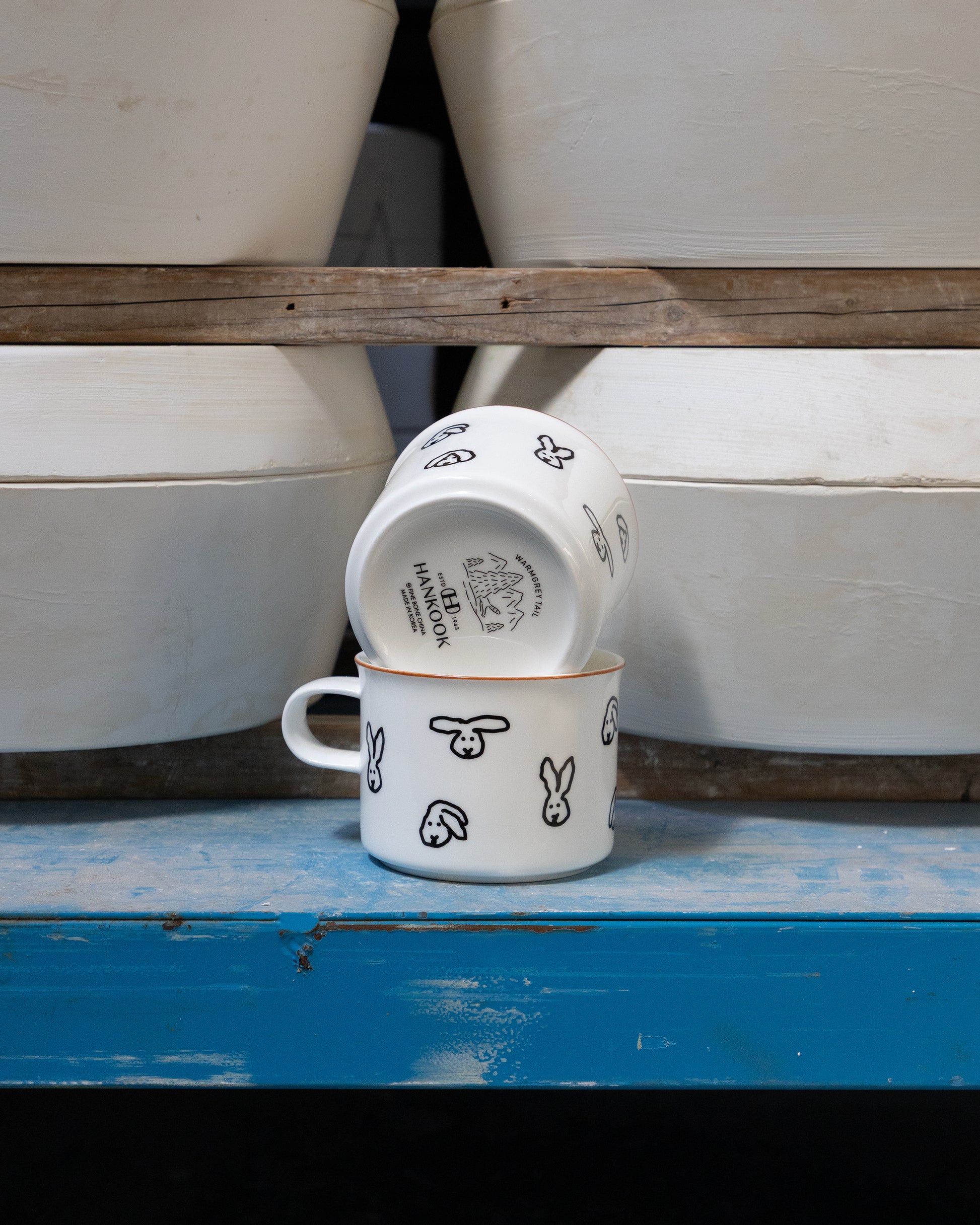 Two white ceramic cups with black designs on a blue surface, with stacked white ceramic pots in the background.
