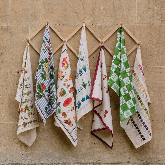 Seven patterned kitchen towels hanging from a wooden diamond rack against a beige stone wall.