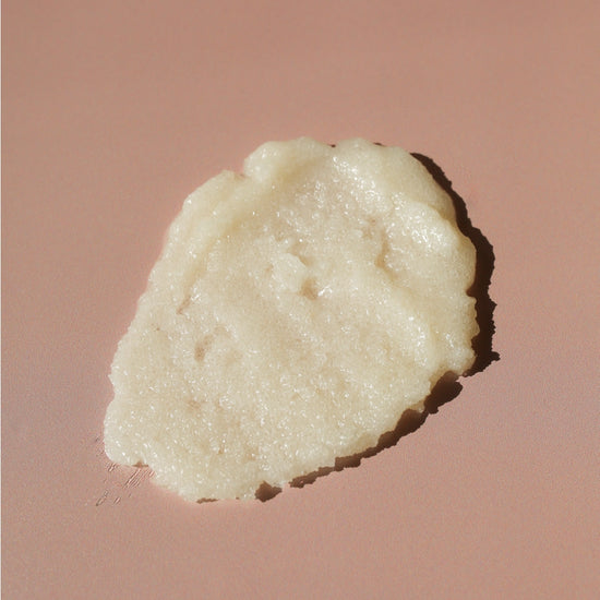 A dollop of creamy, off-white cosmetic cream or balm with a textured, slightly granular surface, placed on a solid dusty rose background.