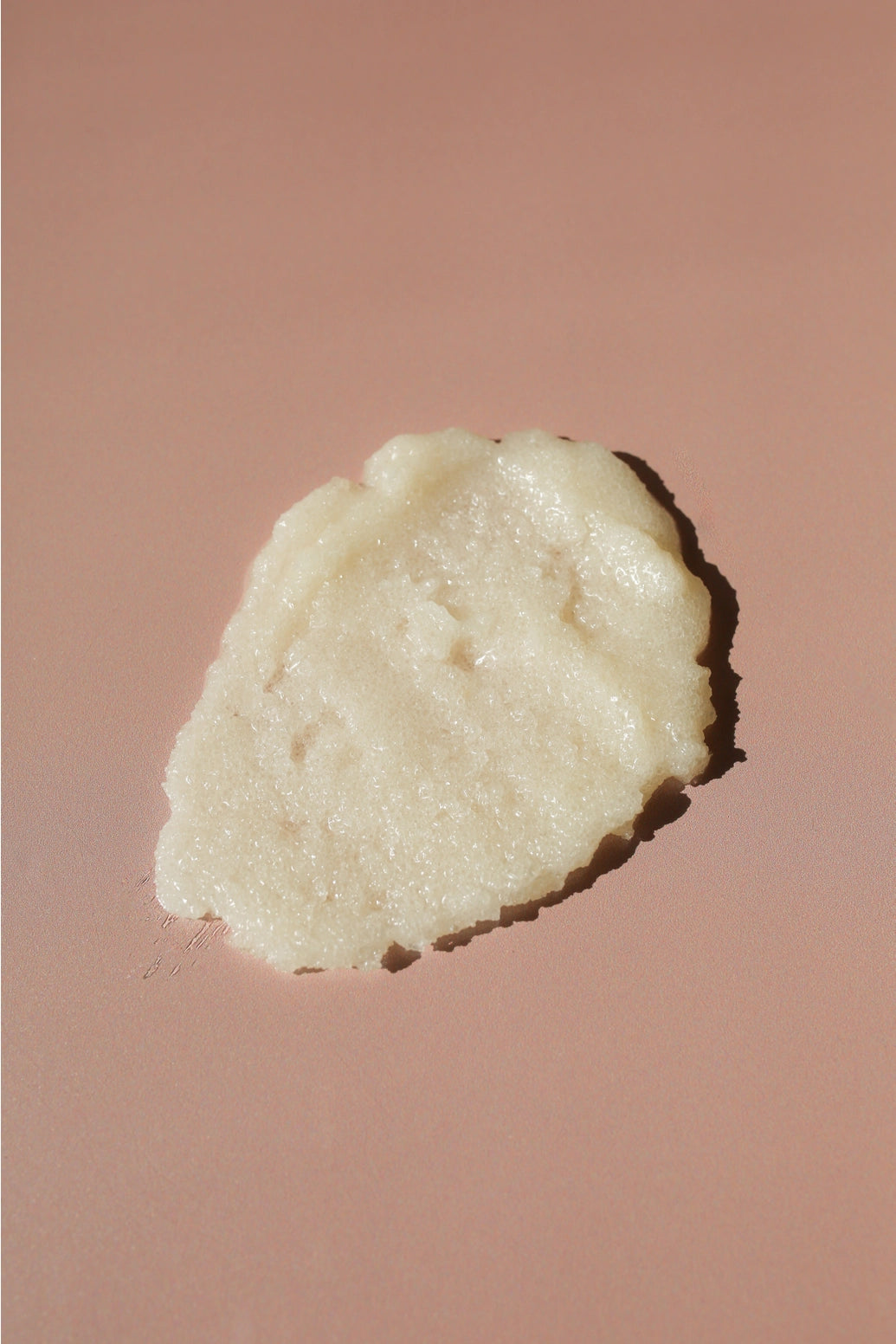 A dollop of creamy, off-white cosmetic cream or balm with a textured, slightly granular surface, placed on a solid dusty rose background.
