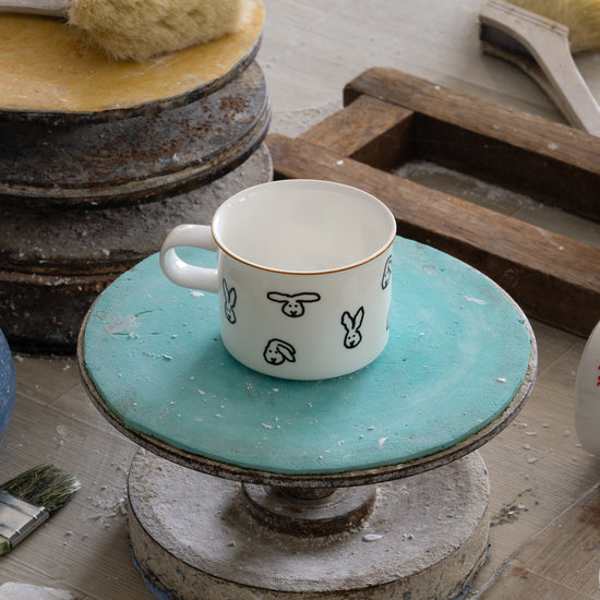 Ceramic mug with rabbit design on a turquoise stand, surrounded by pottery tools and materials.