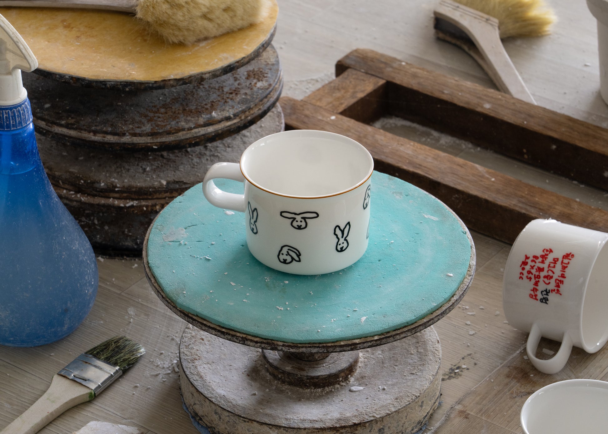 Ceramic mug with rabbit design on a turquoise stand, surrounded by pottery tools and materials.