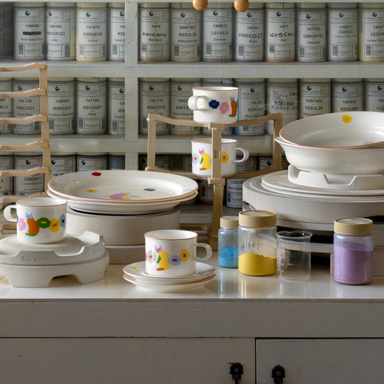 Display of ceramic items with colorful designs on a white surface, with shelves of labeled containers in the background.