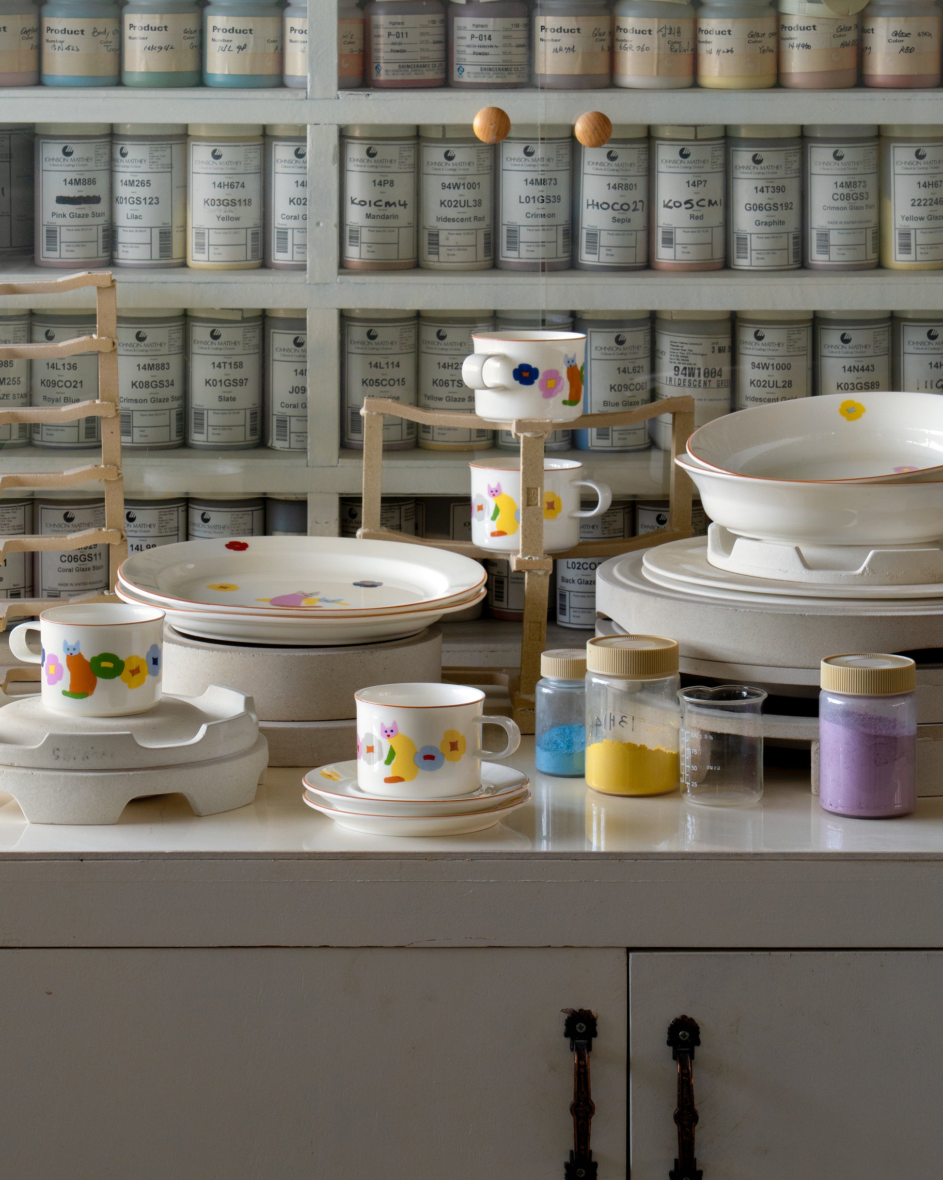 Display of ceramic items with colorful designs on a white surface, with shelves of labeled containers in the background.