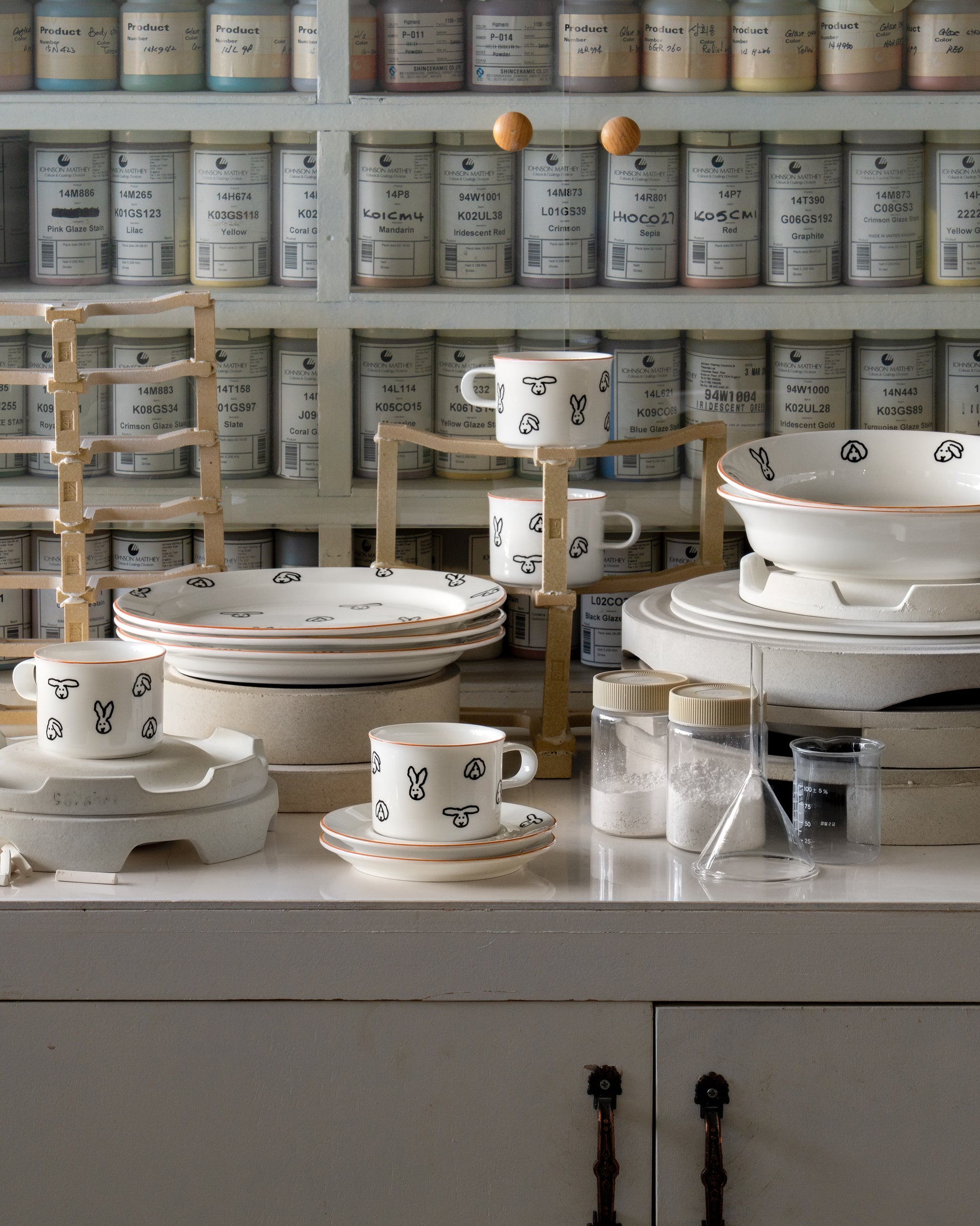 Ceramic dishes with black patterns on a white surface, with shelves of jars in the background.