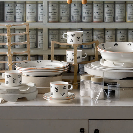 Ceramic dishes with black patterns on a white surface, with shelves of jars in the background.