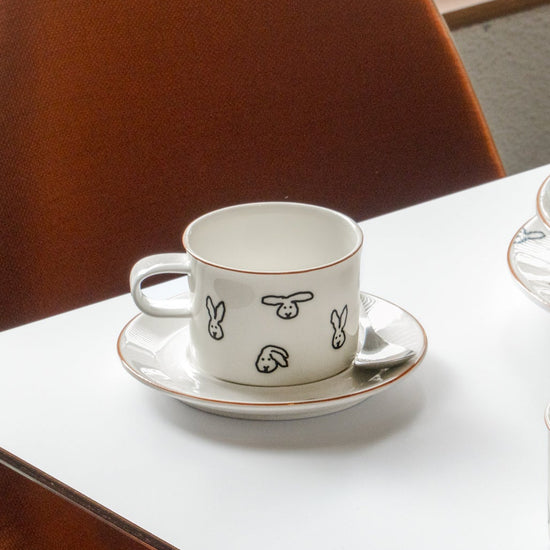 White ceramic cup with black illustrations on a saucer, placed on a white table.