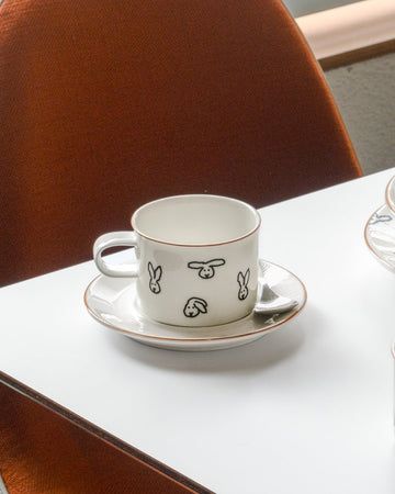 White ceramic cup with black illustrations on a saucer, placed on a white table.