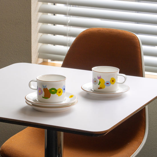 Two ceramic cups with saucers on a small table in front of a window with blinds.