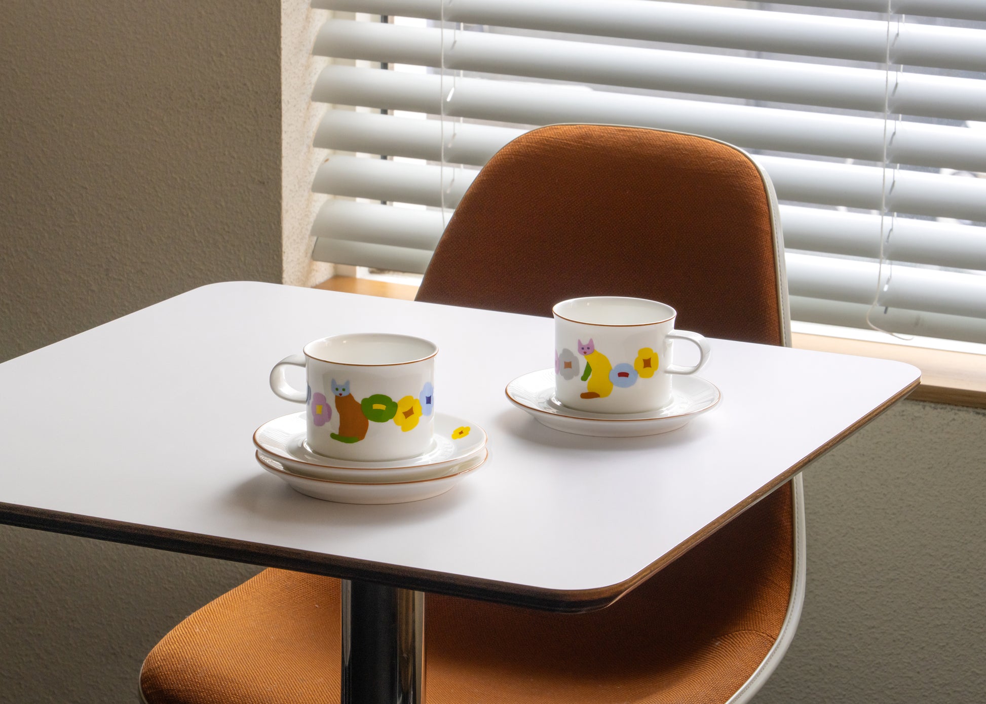 Two ceramic cups with saucers on a small table in front of a window with blinds.