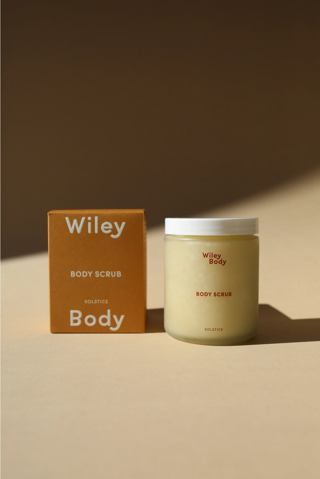 A jar of Wiley Body Scrub next to its brown cardboard packaging box on a warm beige surface with soft shadows.