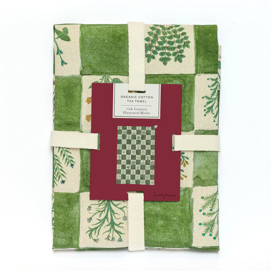 A folded cotton tea towel with a green and cream patchwork botanical print, wrapped with two cream elastic bands and a central red paper label.