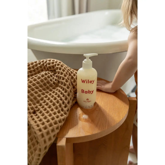 A bottle of Wiley Baby baby shampoo sits on a wooden stool next to a waffle-weave bath towel, with a child standing by a bathtub in the background.