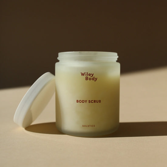 A frosted glass jar of Wiley Body Body Scrub with its white lid leaning against it on a warm beige surface, set against a dark brown background with soft diagonal shadows.