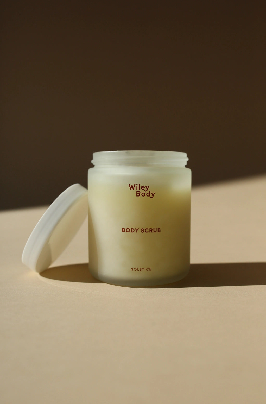 A frosted glass jar of Wiley Body Body Scrub with its white lid leaning against it on a warm beige surface, set against a dark brown background with soft diagonal shadows.