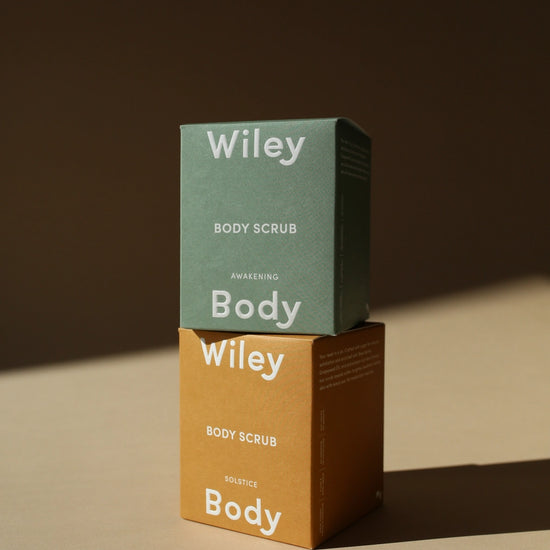 Two stacked Wiley Body Scrub boxes on a warm beige surface against a dark brown background. The top box is sage green and the bottom box is mustard yellow, both with white text reading 'Wiley Body Scrub' and 'Harvesting Body'.