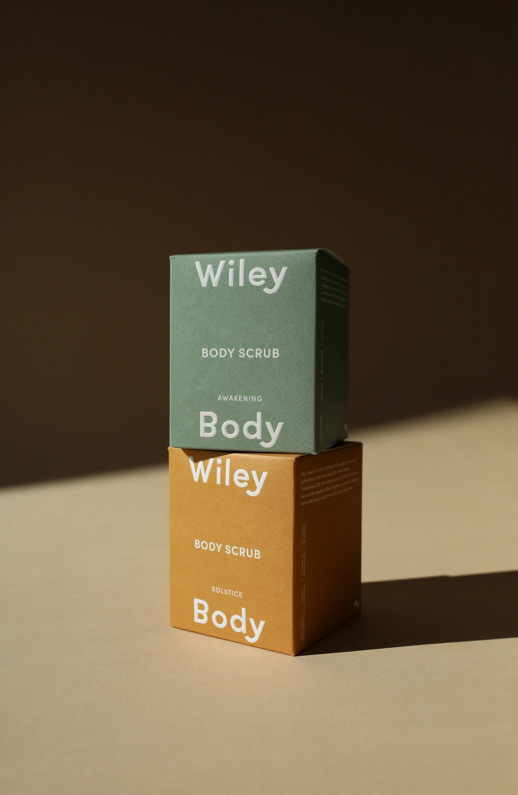 Two stacked Wiley Body Scrub boxes on a warm beige surface against a dark brown background. The top box is sage green and the bottom box is mustard yellow, both with white text reading 'Wiley Body Scrub' and 'Harvesting Body'.