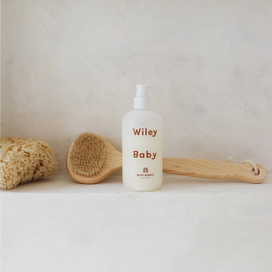 A bottle of Wiley Baby Body Wash with a white pump dispenser, set on a light stone surface with a wooden brush and natural loofah sponge in the background.