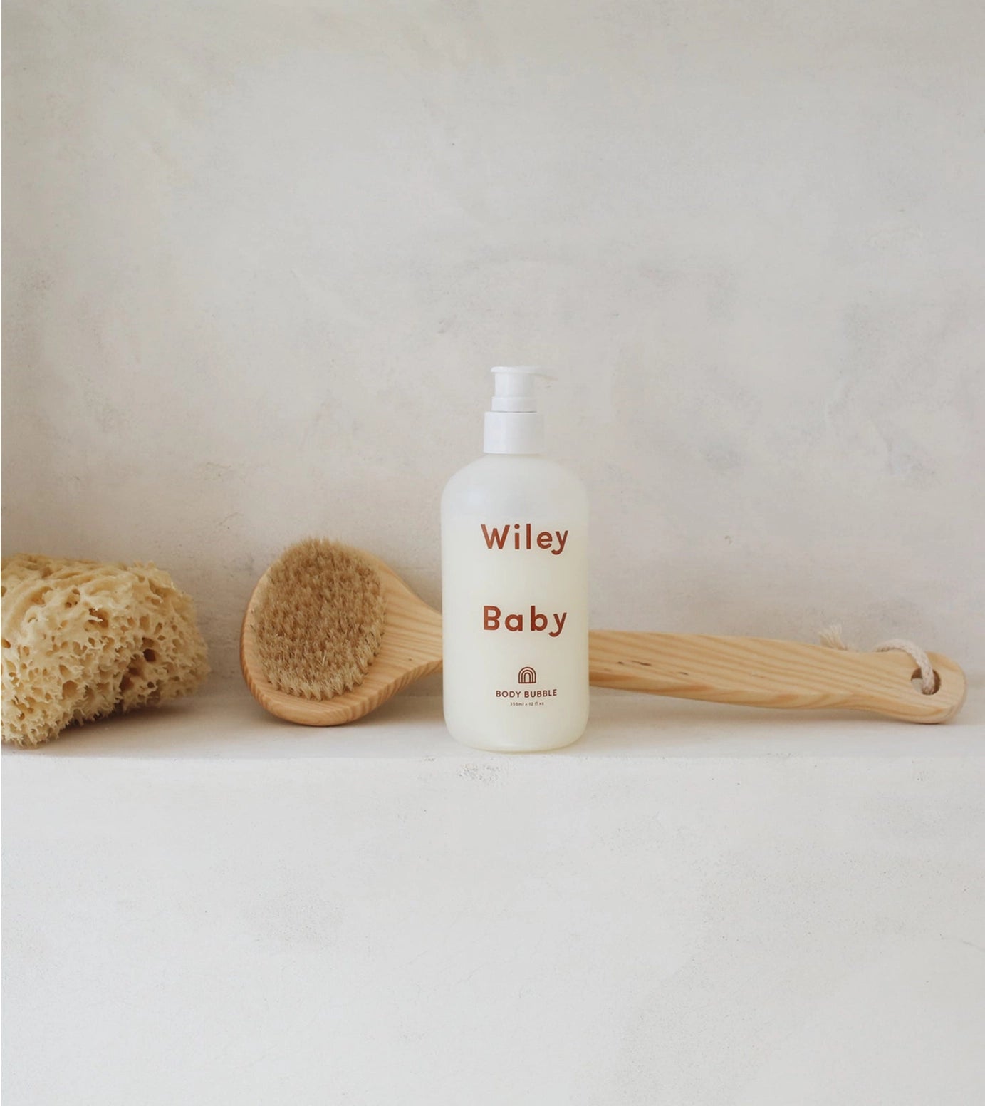 A bottle of Wiley Baby Body Wash with a white pump dispenser, set on a light stone surface with a wooden brush and natural loofah sponge in the background.