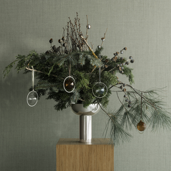 Decorative arrangement with greenery, ornaments, and a metallic vase on a wooden stand against a gray background.