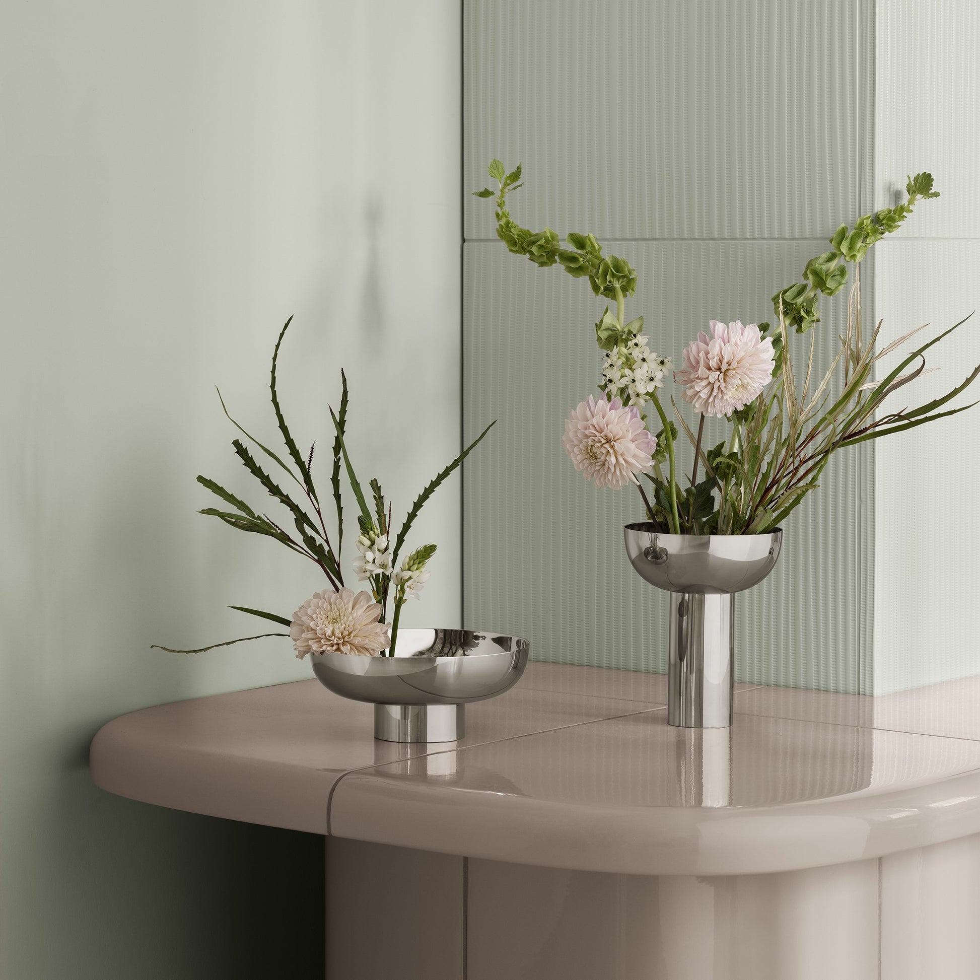 Two silver vases with flowers on a round table against a light green wall.