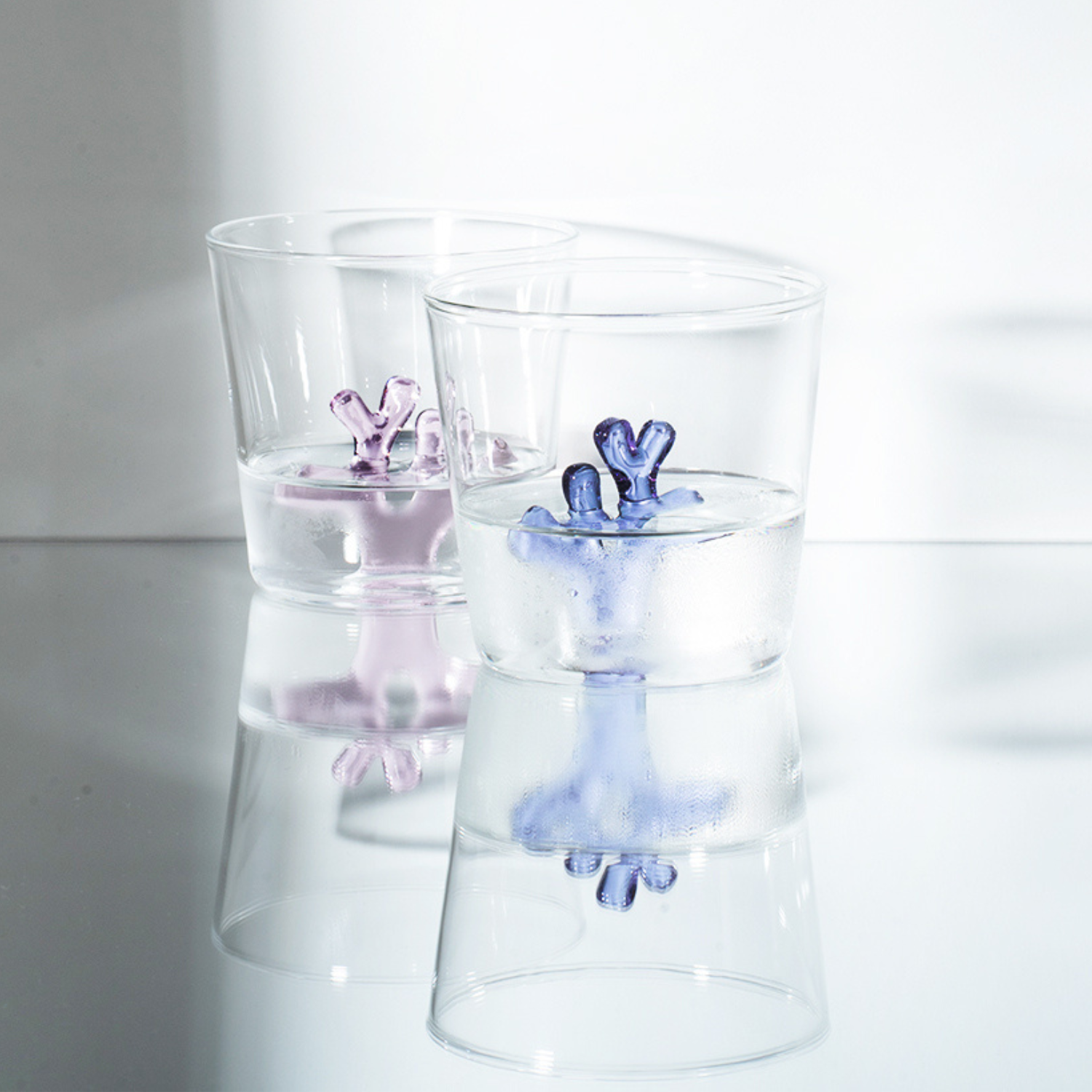 Two clear glass tumblers with pink and blue coral design inside on a reflective surface.