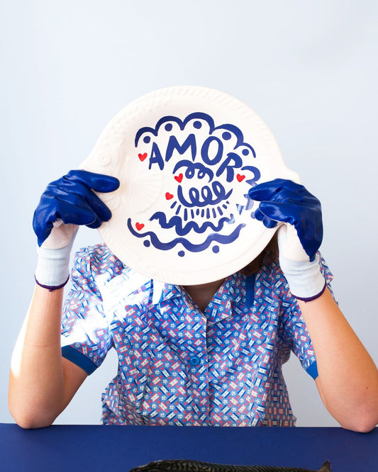 Person wearing gloves holding a decorative plate with 'AMOR' design, sitting at a table with a fish in front.