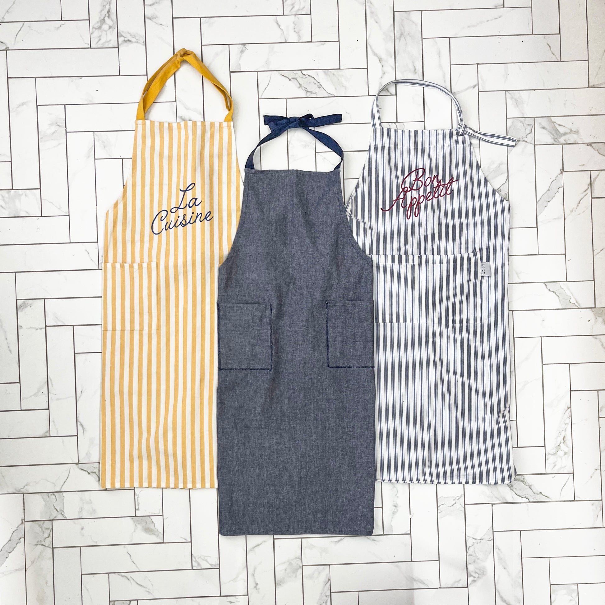 Three aprons with different designs and colors on a marble floor background.