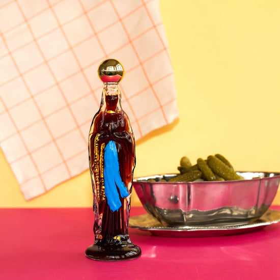 Bottle-shaped container with a blue towel inside, next to a bowl of pickles on a pink surface with a yellow background.