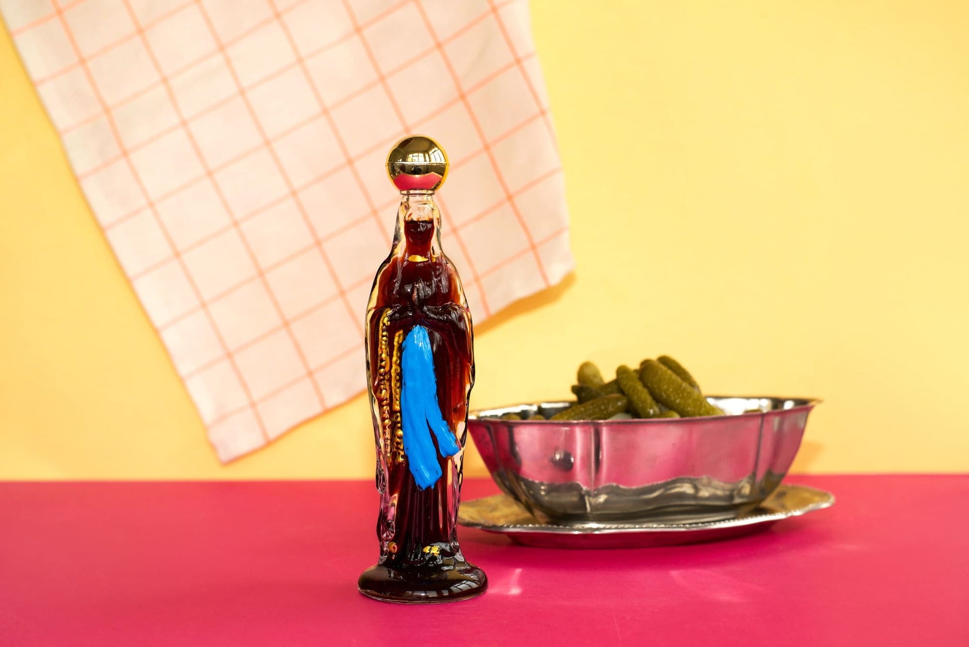 Bottle-shaped container with a blue towel inside, next to a bowl of pickles on a pink surface with a yellow background.