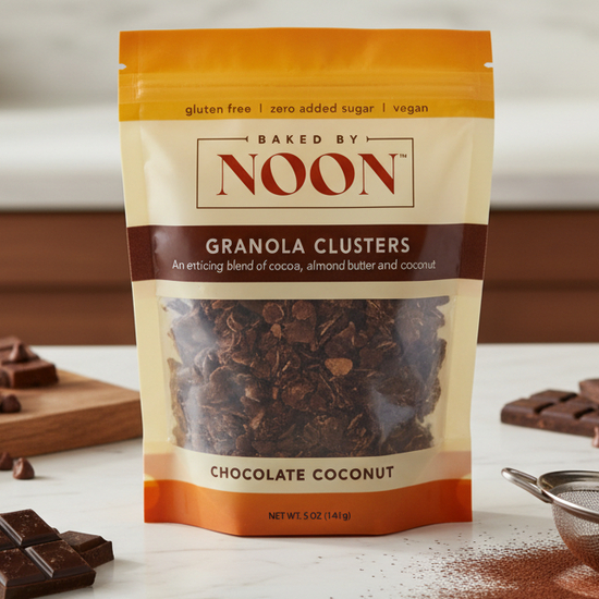 Bag of Baked by Noon Chocolate Coconut Granola Clusters on a white surface with chocolate pieces and a sieve.