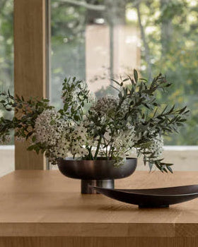 Modern dining area with wooden table, gray chair, and black stool in a room with large windows showing trees.