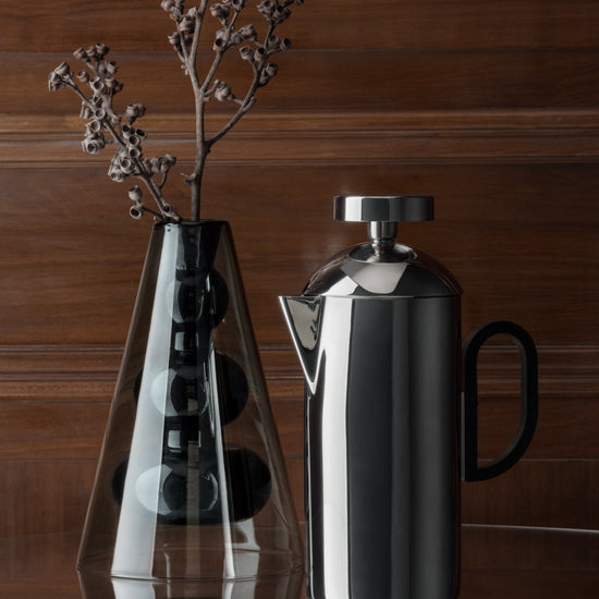 Silver brew cafetiere on a reflective surface with a wooden background