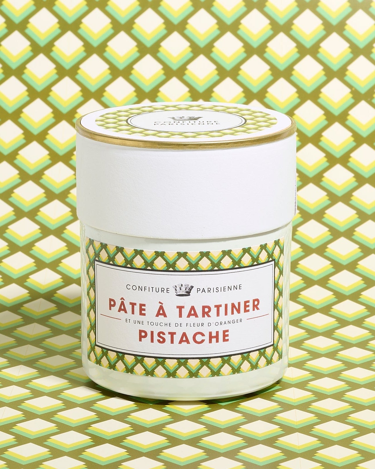 A jar of pistachio spread with a green patterned label, placed against a matching geometric background