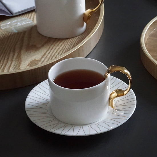 Two white teacups with gold handles on saucers, placed on a dark surface with an open book and wooden trays.