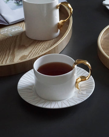 Two white teacups with gold handles on saucers, placed on a dark surface with an open book and wooden trays.