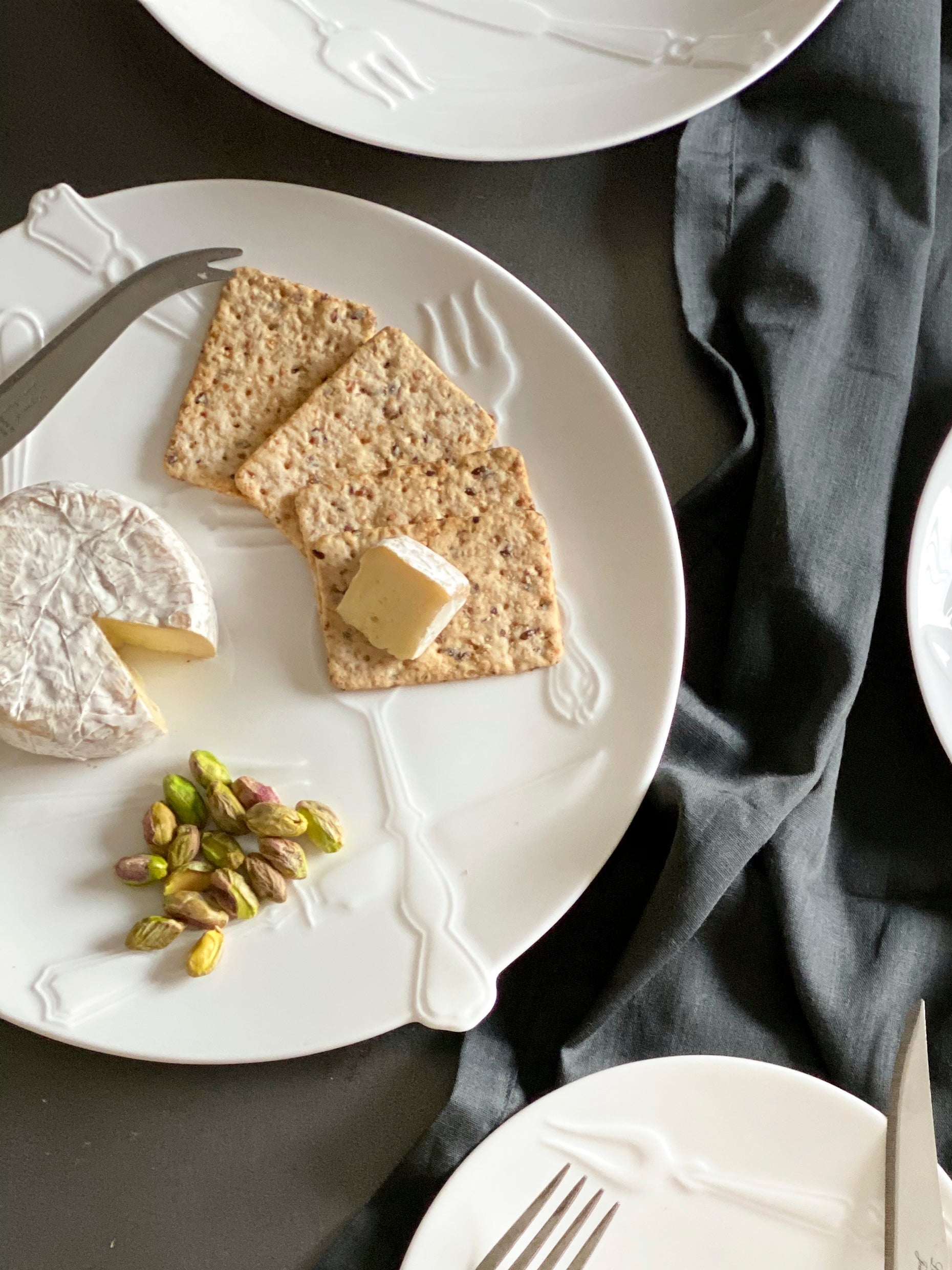 Dessert setting with plates of cake, tart, and cheese on a dark fabric surface.