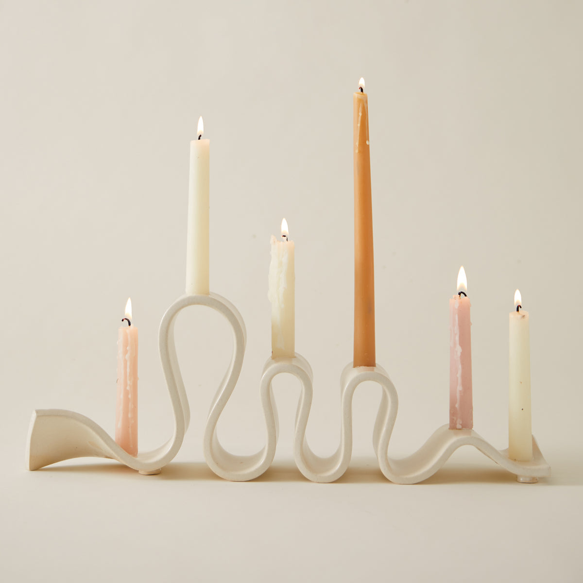 Cream ceramic wavy candlestick holder with six lit taper candles in soft pastel and neutral tones, set against a neutral background