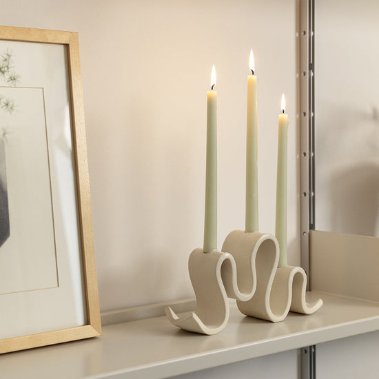 Cream-colored ceramic candelabra with three curved arms, holding three unlit taper candles, set against a plain neutral background