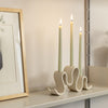 Cream-colored ceramic candelabra with three curved arms, holding three unlit taper candles, set against a plain neutral background