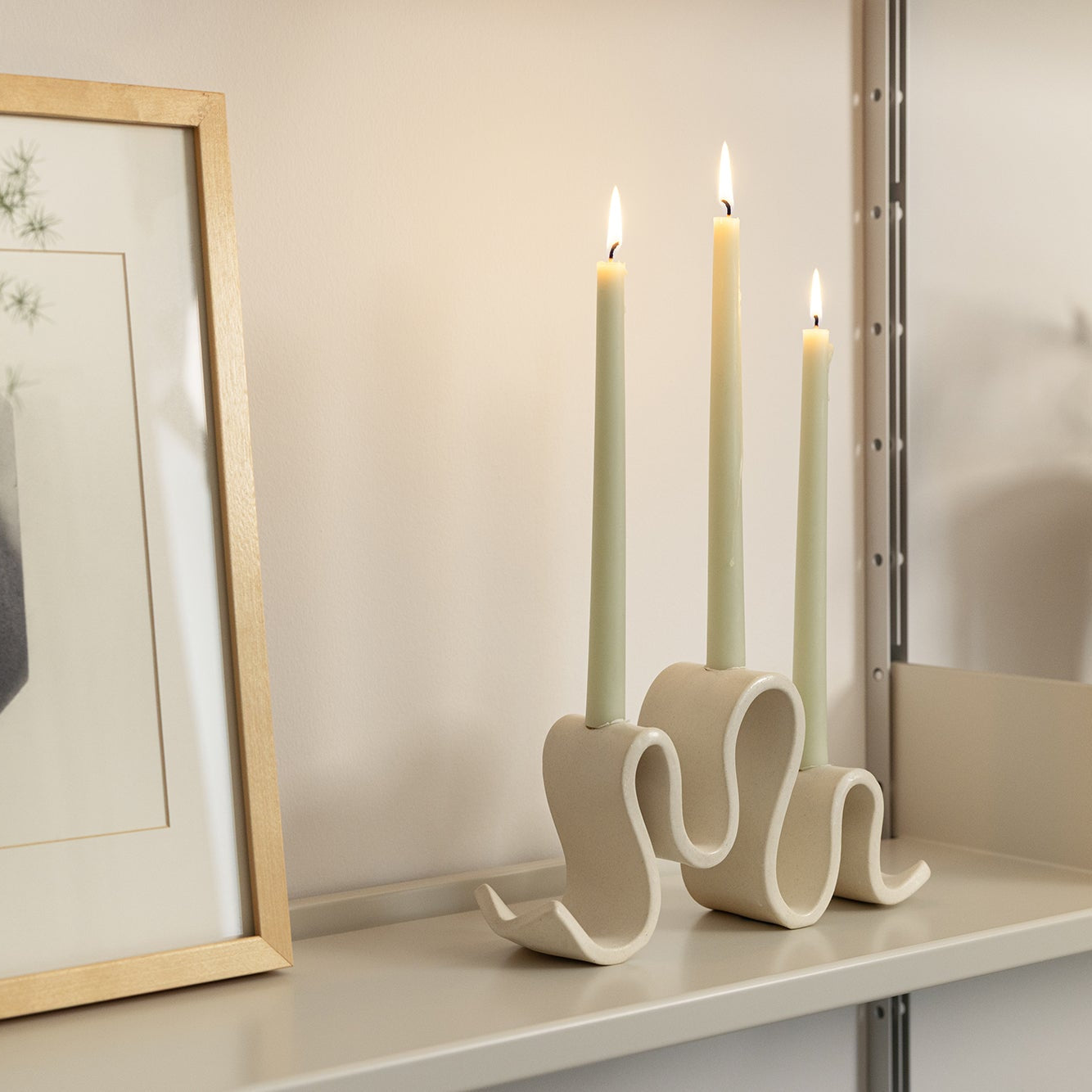 Cream-colored ceramic candelabra with three curved arms, holding three unlit taper candles, set against a plain neutral background