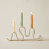 Cream-colored ceramic candelabra with a wavy, sculptural base holding three lit taper candles against a plain neutral background