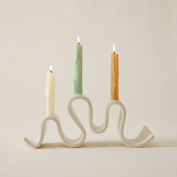 Cream-colored ceramic candelabra with a wavy, sculptural base holding three lit taper candles against a plain neutral background