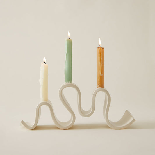 Cream-colored ceramic candelabra with a wavy, sculptural base holding three lit taper candles against a plain neutral background