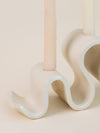 Close up of a cream-colored ceramic candelabra with a wavy base holding three unlit taper candles, shown against a plain neutral background.