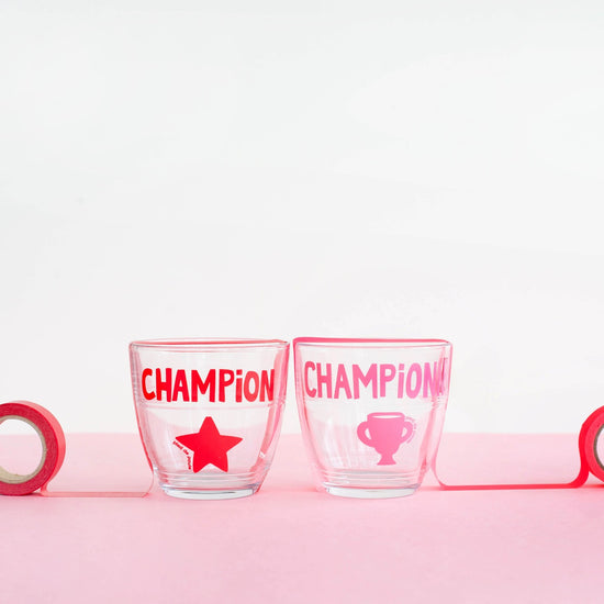 Two glass tumblers with 'CHAMPION' and star design on a pink surface.