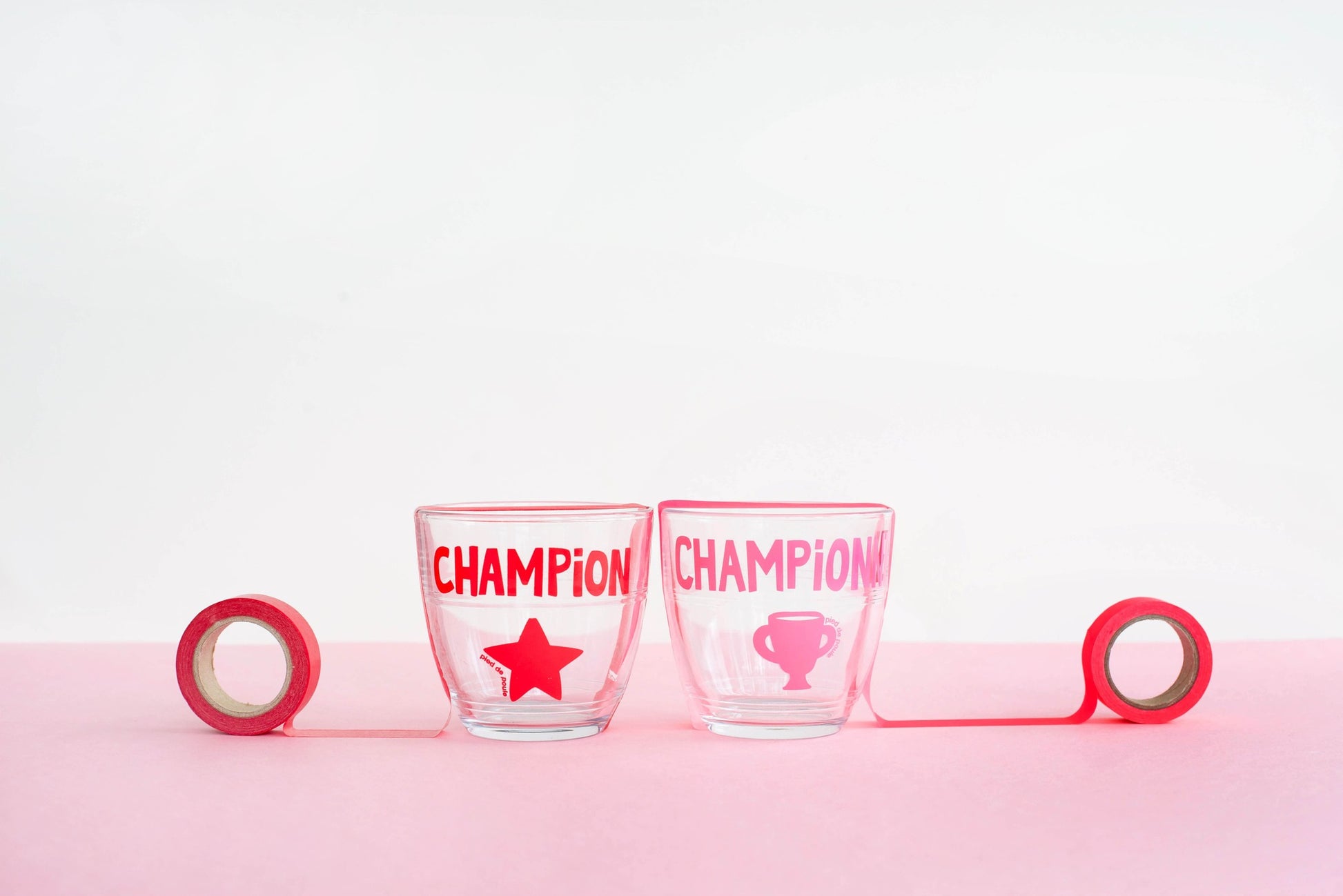Two glass tumblers with 'CHAMPION' and star design on a pink surface.