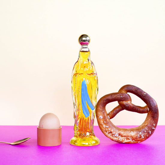 Bottle with a colorful design, a pretzel, an egg, and a spoon on a pink surface with a light purple background.