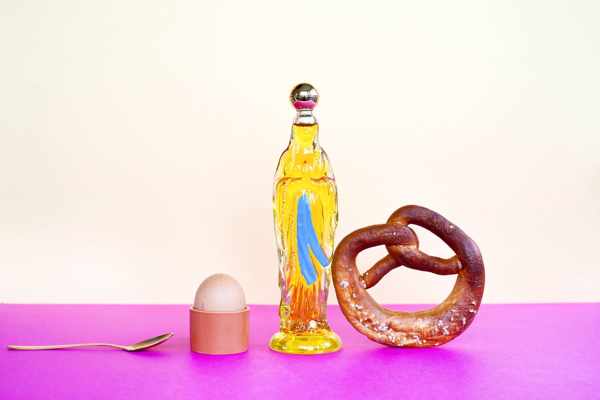 Bottle with a colorful design, a pretzel, an egg, and a spoon on a pink surface with a light purple background.